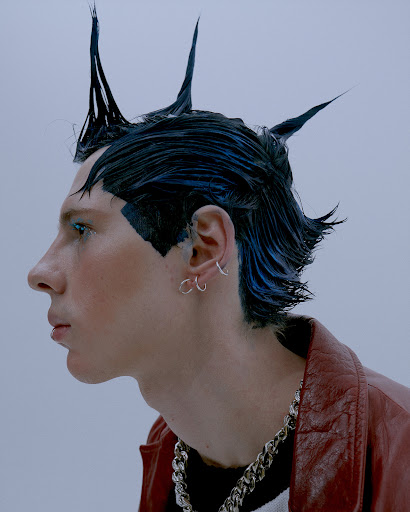 Close-up studio shot of an adult with a punk hairstyle, wearing a red jacket and silver chain necklace against a light grey background.