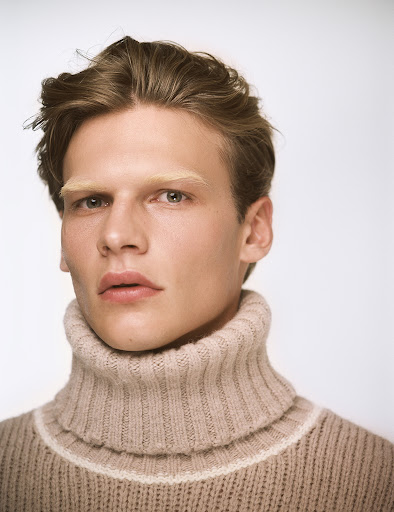 Close-up portrait of a fair-skinned young adult male with blond hair, wearing a beige turtleneck sweater against a light backdrop.