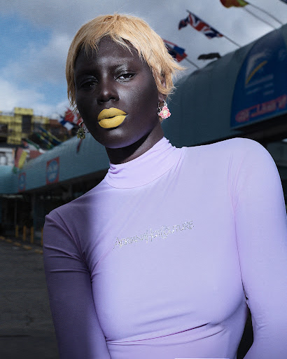 Dark-skinned woman with blonde hair and yellow lipstick poses in lavender top outdoors with flags and architectural structures visible.