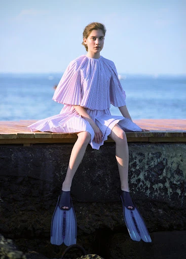 Young woman in lavender dress and flippers sits on dock with ocean background in a fashion editorial shot during the daytime.