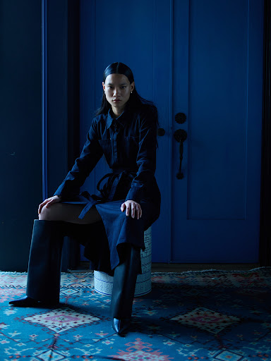 Portrait of a woman with dark hair in a navy belted dress and black boots, sitting on a stool against a blue door and patterned rug.