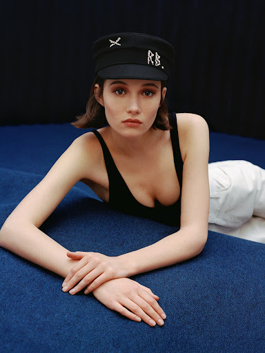 Portrait of a young woman lying on blue fabric, wearing a black top and cap with white trousers, indoors with a dark background.
