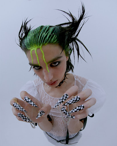 Model poses with bold makeup, green-streaked hair, a spiked choker, and long, checkered-patterned nails against a plain background.