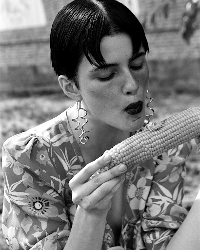 Black and white portrait of a woman with short dark hair, eating corn on the cob. She's wearing a floral shirt and statement earrings.