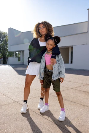 Two girls, one adult and one child, pose together outdoors in casual fashion wear with a modern building in the background on a sunny day.