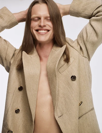 Smiling man with long brown hair poses in a beige blazer with hands behind his head against a white background in a studio shot.
