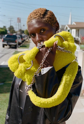 Fashion portrait of a Black woman wearing a yellow faux fur bag with a silver chain, standing on a residential street on a sunny day.