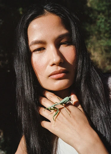 Close-up portrait of a woman with long black hair, holding a green praying mantis on her hand in an outdoor setting with green foliage.