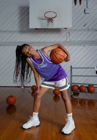 Young girl with braided hair posing in a basketball jersey and shorts holding a basketball inside a gym with wood floors