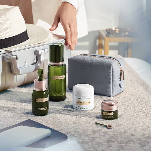 La Mer skincare products sit on a patterned surface near a silver suitcase and gray cosmetic bag, with a woman's hand visible in the background.