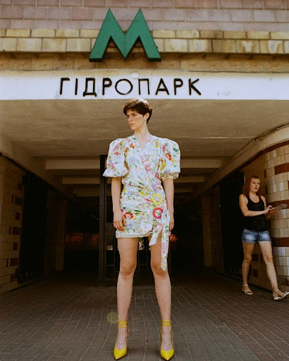 A woman in a floral dress and yellow heels stands in front of a metro entrance, with another person in the background.