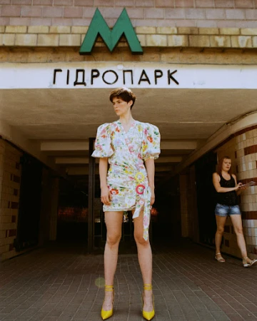 A woman in a floral dress and yellow heels stands in front of a metro entrance, with another person in the background.