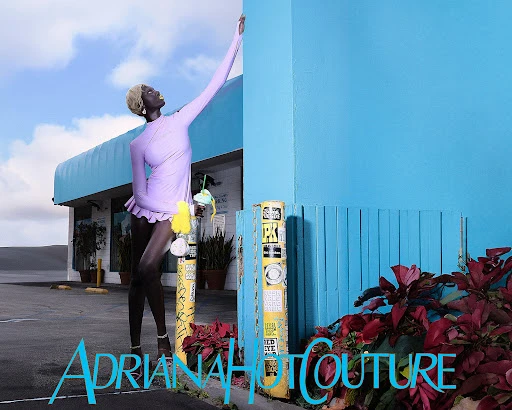 Fashion shot of a slender model in a lavender dress with blonde hair, posing against a turquoise building and blue sky with clouds.