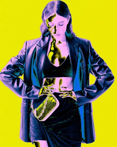 A woman in a black crop top and skirt, with a blue blazer and a small bag, poses in front of a yellow background in a fashion style image.