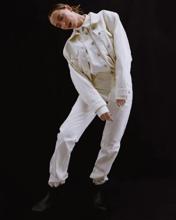 Model in white denim jacket and trousers posing against a dark background, styled by Mari Siviakova.