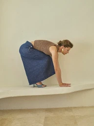 Woman in brown knit top and blue denim skirt crouches on a low white structure styled by Mari Siviakova