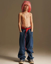 A young model standing shirtless wearing baggy blue jeans and a red belt with a pink draped head covering.