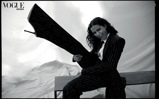 Monochrome fashion editorial image featuring a smiling person holding a giant stiletto shoe while wearing a pinstripe suit