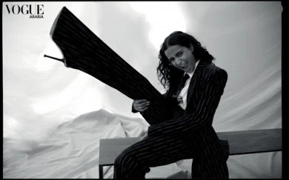 Monochrome fashion editorial image featuring a smiling person holding a giant stiletto shoe while wearing a pinstripe suit