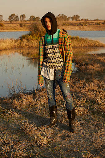 Fashion portrait of a person outdoors. They are wearing a hooded shirt, plaid jacket, blue jeans and black cowboy boots in a nature setting.