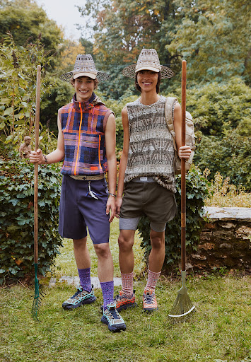 Two smiling adult men model sleeveless shirts, shorts, and matching hats, holding rakes in a lush green garden setting.