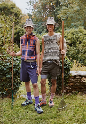 Two smiling adult men model sleeveless shirts, shorts, and matching hats, holding rakes in a lush green garden setting.