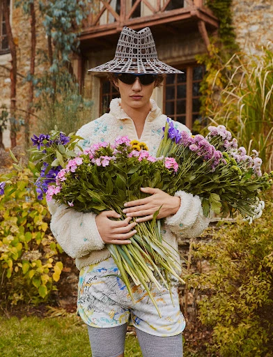 A person holding colorful flowers, wearing sunglasses and a hat, with a textured top and shorts, standing outdoors near a stone building.