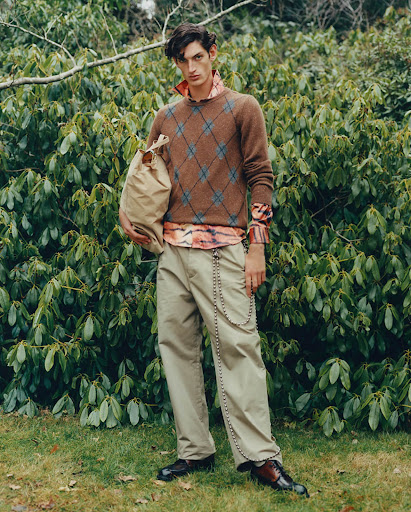 A male fashion model poses outdoors wearing a brown argyle sweater, khaki trousers with chain, and leather shoes while holding a bag.