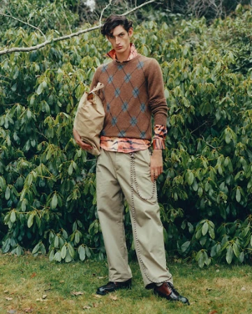 A male fashion model poses outdoors wearing a brown argyle sweater, khaki trousers with chain, and leather shoes while holding a bag.