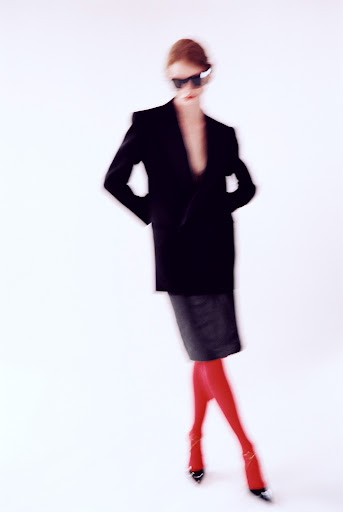 Fashion portrait of a woman in a black blazer, skirt, and red tights, posing against a bright white background with a blurred effect.