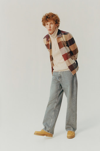 Full shot of a red-haired male model in a multicolored flannel shirt, light turtleneck, wide jeans, and loafers, posing in a photo studio.