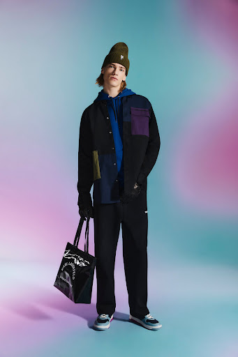 Full-length studio shot of a young adult male fashion model in a color-blocked shirt, black pants, beanie, holding a glossy black tote bag.
