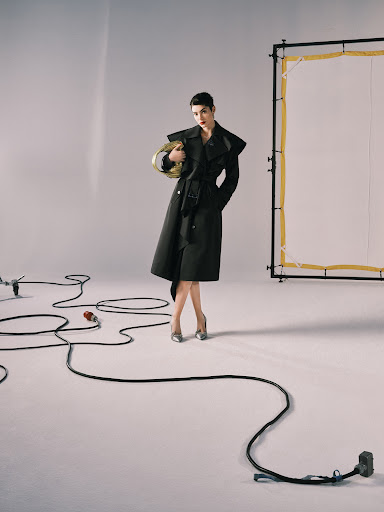 A stylish woman in a black trench coat poses in a photo studio, holding a golden headdress against a neutral backdrop with studio equipment.