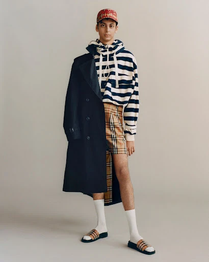 Man in striped hoodie, plaid shorts, socks and slides stands with coat draped over shoulder against neutral studio backdrop.