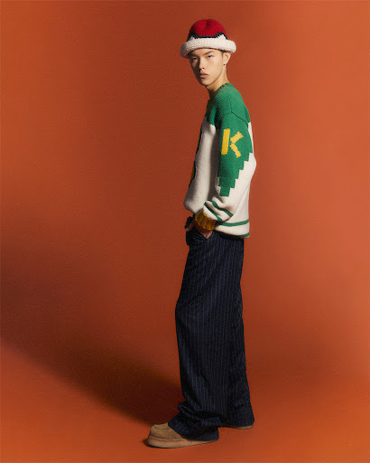 A stylish young Asian male model posing in a studio wearing a knit hat, graphic sweater, and pinstripe pants against a solid orange backdrop.