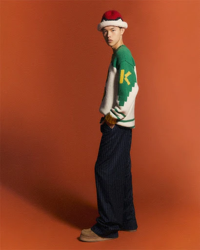 A stylish young Asian male model posing in a studio wearing a knit hat, graphic sweater, and pinstripe pants against a solid orange backdrop.