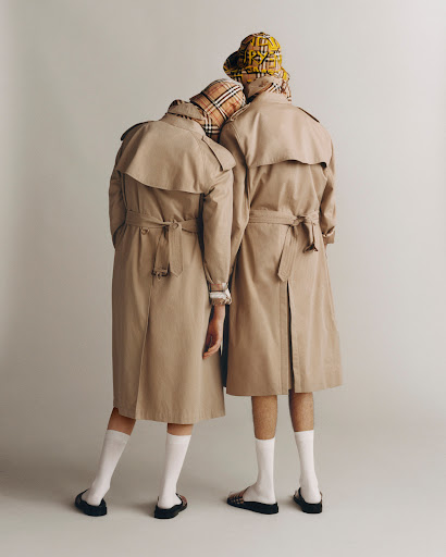 Two people in tan trench coats and patterned bucket hats stand against a light grey background, facing away from the camera.