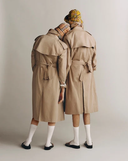 Two people in tan trench coats and patterned bucket hats stand against a light grey background, facing away from the camera.