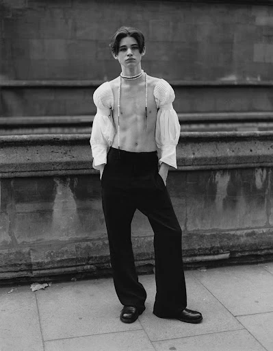 A young adult stands confidently in high-waisted trousers and a voluminous short-sleeved top, accessorized with a choker and pearl necklace.
