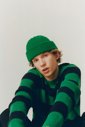 Portrait of an adult man wearing a green beanie and a green and black striped sweater, posing indoors against a white backdrop.