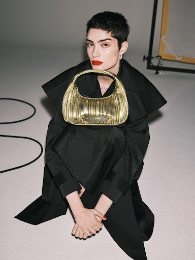 Adult female with short, dark hair sits on white floor holding a gold handbag, in a fashion studio with lighting equipment in the background.