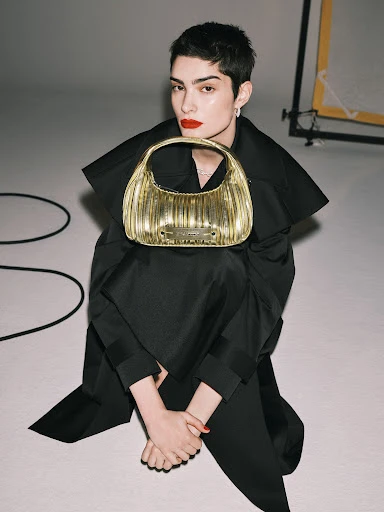Adult female with short, dark hair sits on white floor holding a gold handbag, in a fashion studio with lighting equipment in the background.