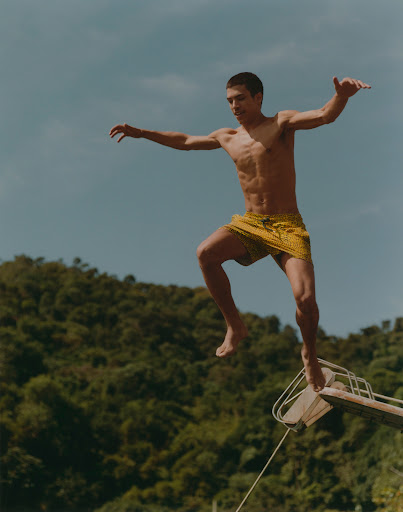 Athletic man in mid-air performs a jump from a platform against a background of a lush green hillside and a blue sky on a sunny day.