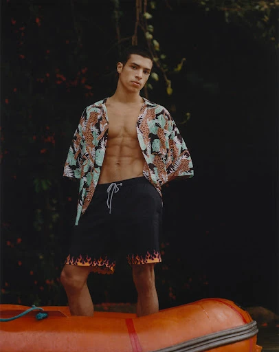 A young adult male stands in an orange inflatable boat, wearing a printed short-sleeve shirt and swim trunks with a flame design.