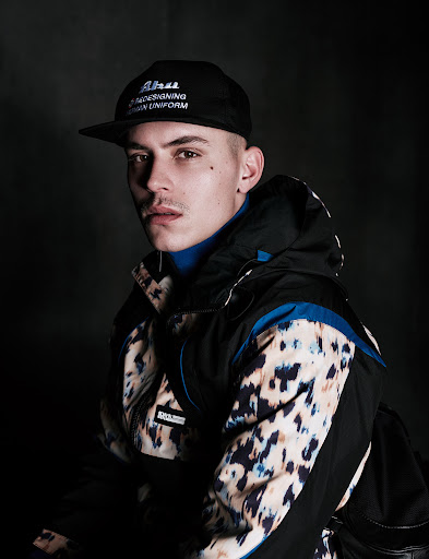 Portrait of a young man wearing a black baseball cap, a blue turtleneck, and a patterned black-and-tan ski jacket against a dark background.