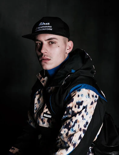 Portrait of a young man wearing a black baseball cap, a blue turtleneck, and a patterned black-and-tan ski jacket against a dark background.