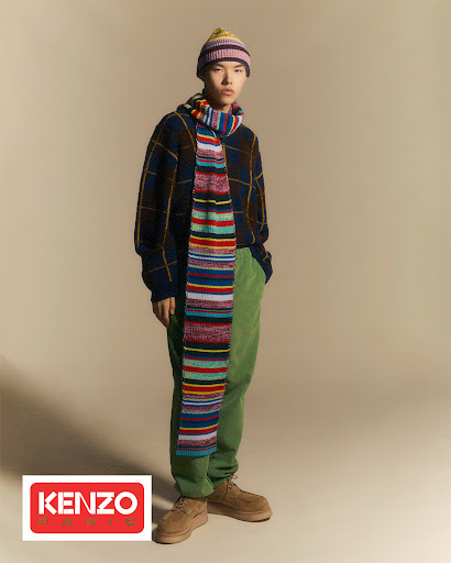 A stylish Asian male model poses in a photo studio wearing a colorful knit hat and scarf with a plaid sweater and green corduroy pants.
