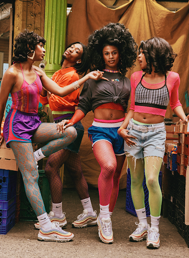 Group of four Black women in 80s-inspired athletic outfits and sneakers pose in urban setting with vintage crates and beige walls.