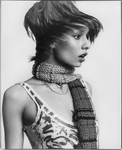 Monochrome portrait of a woman with short hair and a scarf, looking off to the side against a neutral background in a studio setting.