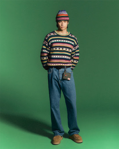 Full-length shot of a young adult man standing in front of a green backdrop, wearing jeans, a sweater, beanie, and loafers.